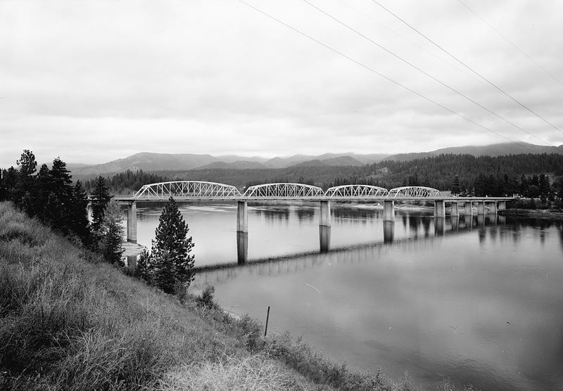 Black-and-white photo of the steel-arch Oldtown Bridge crossing the wide Pend Oreille River, with forested hills behind