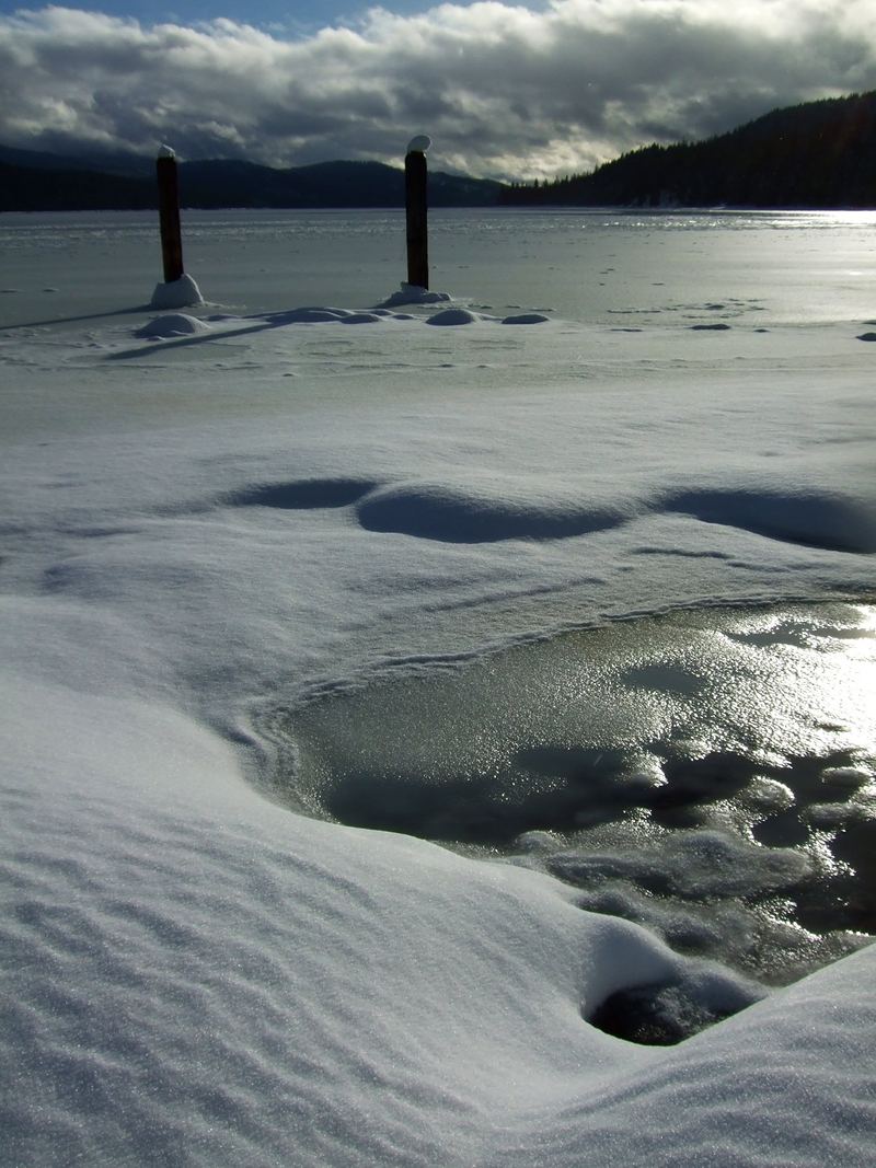 Two old pilings in a snow-covered frozen lake, with mountains under a cloudy sky